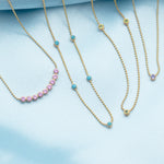 A Birthstone Arc Layering Necklace (Pink Tourmaline), Quintet Birthstone Layering Necklace (Turquoise), Triple Birthstone Layering Necklace (Peridot) and  Single Birthstone Layering Necklace (Tanzanite), all in yellow gold, against a light blue fabric background. 