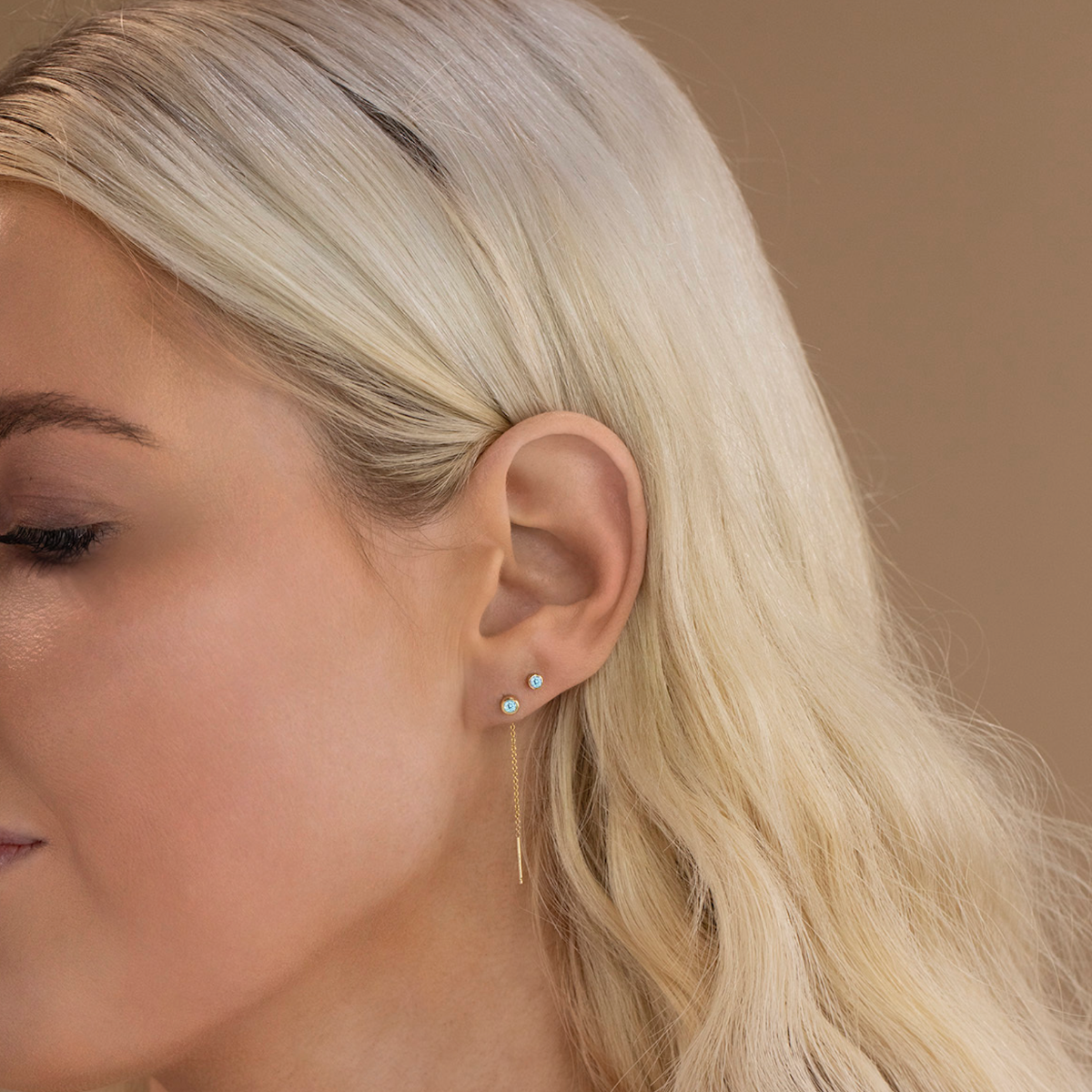 Close-up of a Birthstone Stud and Threader Earring in Aquamarine (March) in a blonde woman's ear against a light brown background. 
