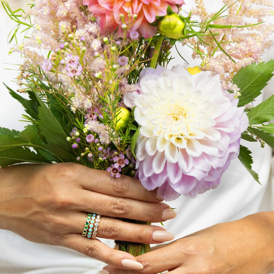 A pair of hands wearing The Fern Bouquet Ring, The Bluebell Bouquet Ring and The Evermore Ring, holding a bunch of flowers. 