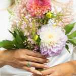 A pair of hands wearing The Fern Bouquet Ring, The Bluebell Bouquet Ring and The Evermore Ring, holding a bunch of flowers. 