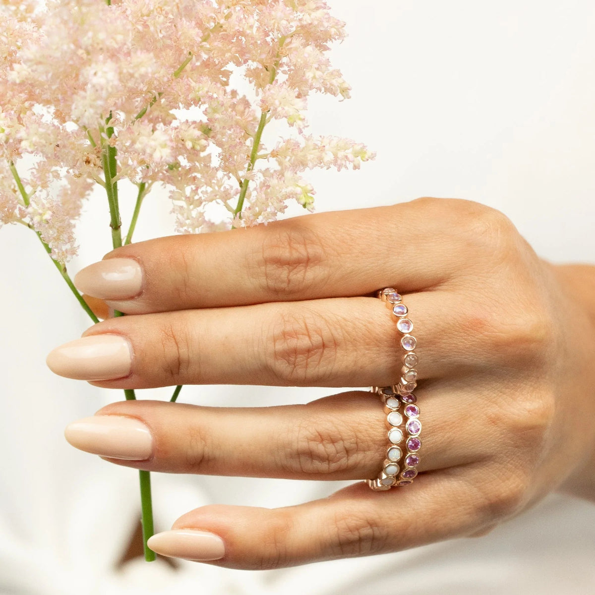 A hand wearing The Peony Bouquet Ring, The Magnolia Bouquet Ring and The Gardenia Bouquet Ring with nude nails holding flowers against a light grey background. 