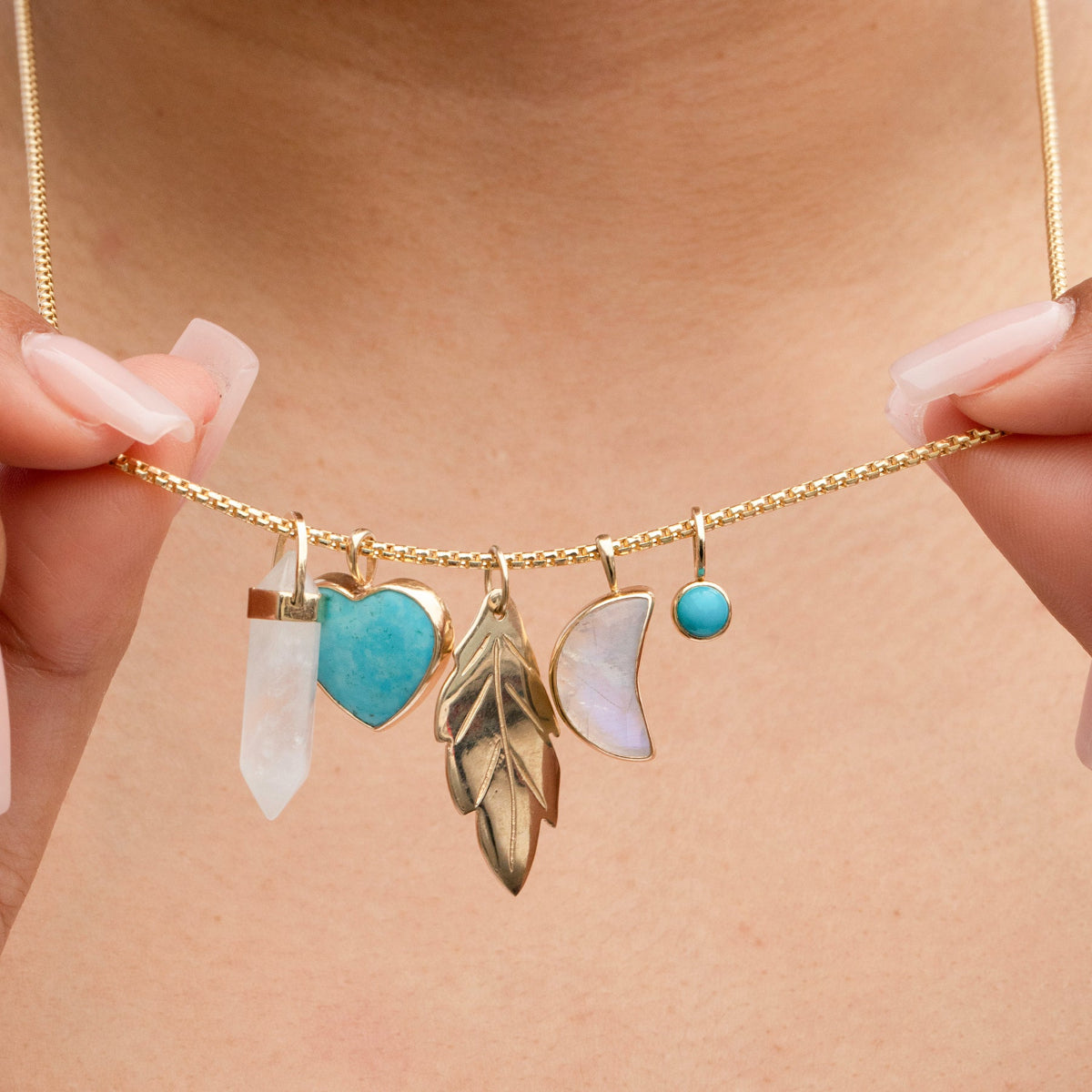 A hand holding a yellow gold Box Chain with several charms, including a Balance Crystal Charm, Turquoise Heart Charm, Moonstone Crescent Charm, 20th Anniversary Leaf Pendant and a Turquoise Dangle Charm. 