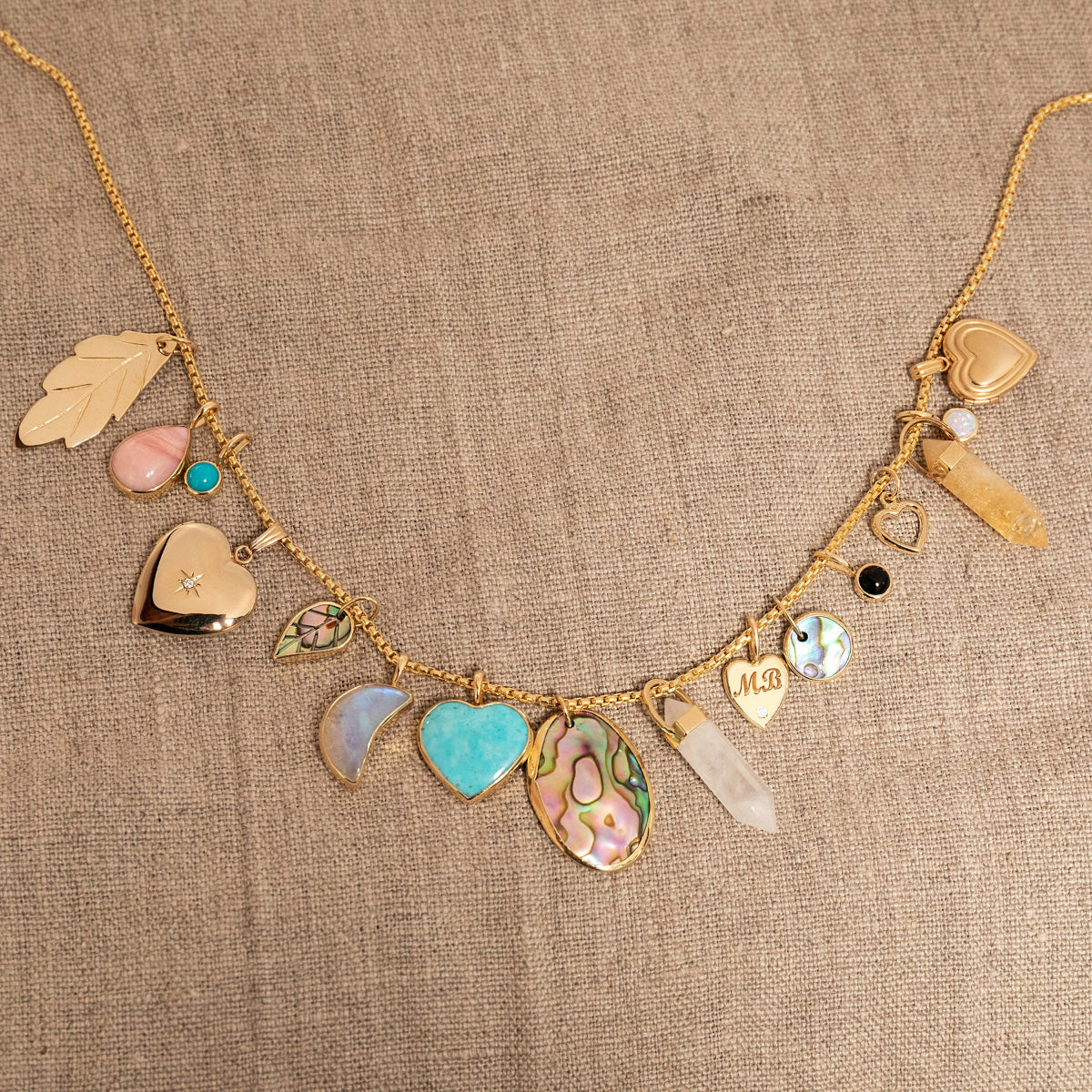 A Box Chain with the following charms - 
20th Anniversary Leaf Pendant, Pink Opal Tear Drop Charm, Turquoise Dangle Charm, Diamond Heart Locket Charm, Abalone Leaf Charm, Moonstone Crescent Charm, Turquoise Heart Charm, Large Abalone Cabochon Charm, two Balance Crystal Charms, Engraved Heart Charm, Small Abalone Cabochon Charm, Onyx Dangle Charm, Cut Out Heart Charm, Opal Dangle Charm and a Heart Locket Charm, against a beige fabric background.