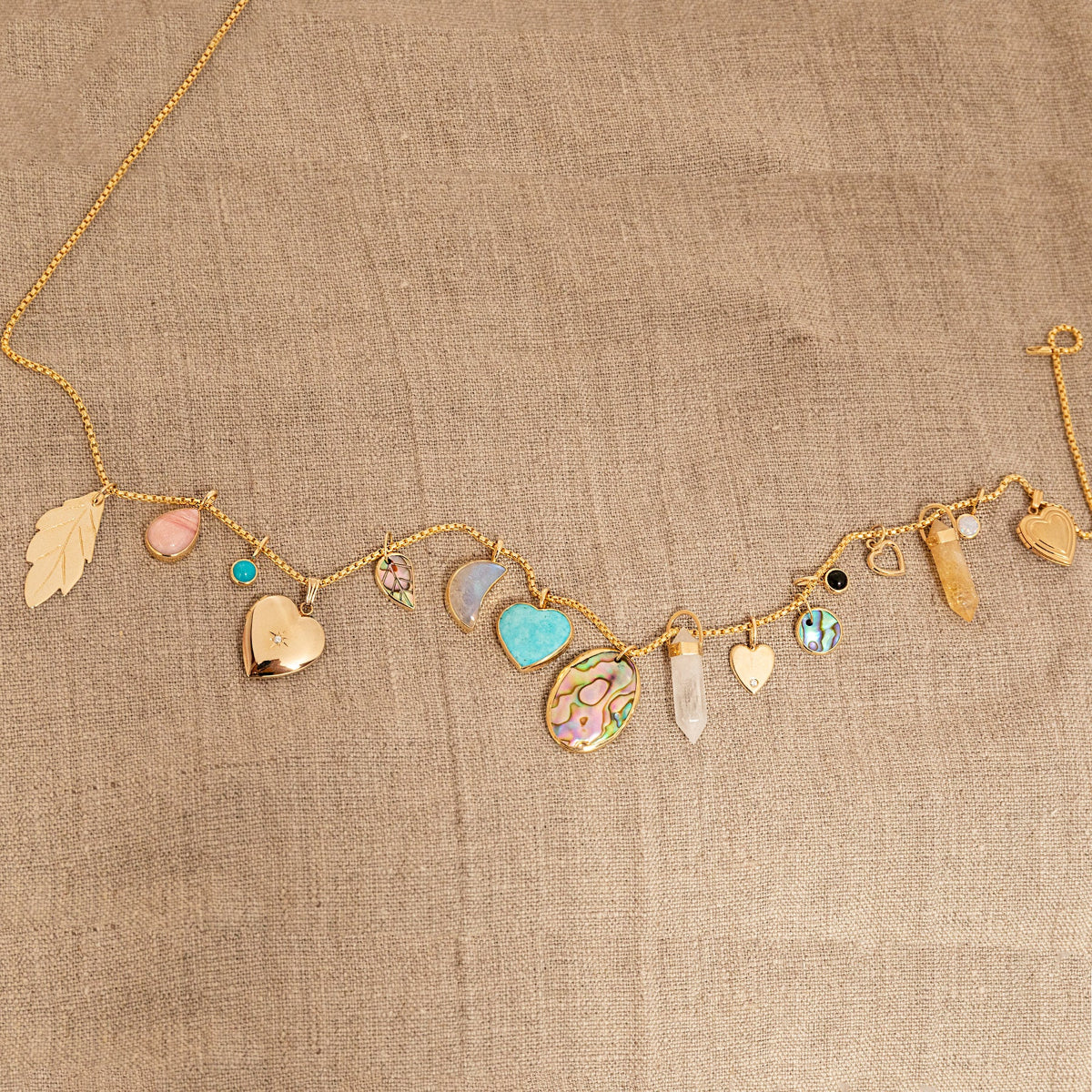 A Box Chain with the following charms - 
20th Anniversary Leaf Pendant, Pink Opal Tear Drop Charm, Turquoise Dangle Charm, Diamond Heart Locket Charm, Abalone Leaf Charm, Moonstone Crescent Charm, Turquoise Heart Charm, Large Abalone Cabochon Charm, two Balance Crystal Charms, Engraved Heart Charm, Small Abalone Cabochon Charm, Onyx Dangle Charm, Cut Out Heart Charm, Opal Dangle Charm and a Heart Locket Charm, against a beige fabric background.