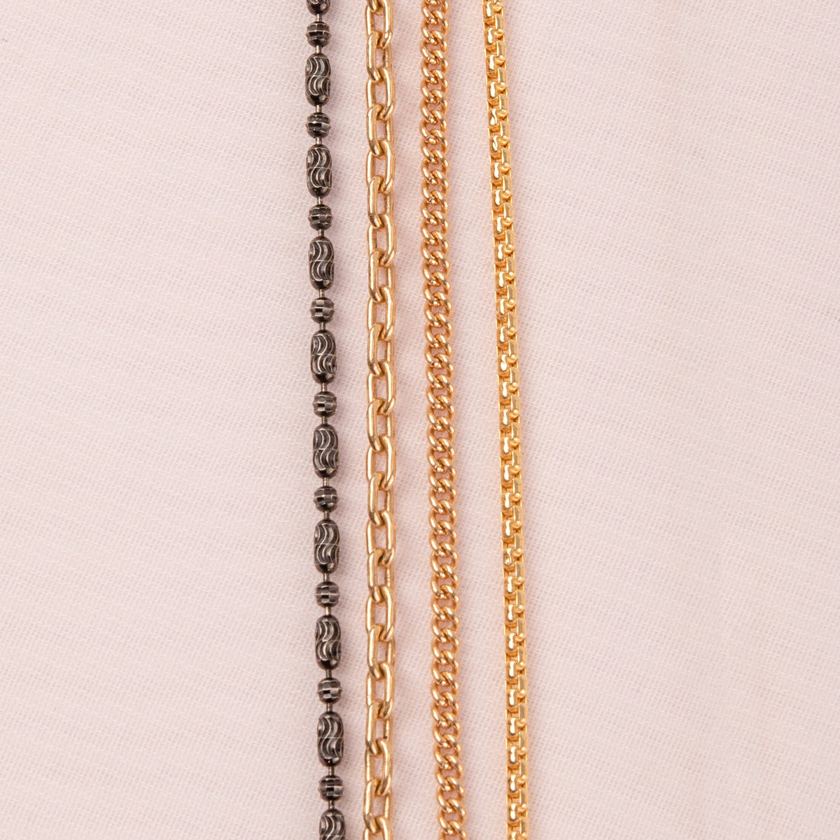 A Black Rhodium Antique Bead Chain, a Round Curb Chain, a Box Chain and a Oval Link Chain laid on an off-white fabric background.