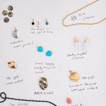 Collection of various jewellery items on white paper with labels, including a yellow Box Chain, Clarity Crystal Charms, Etched Turquoise Heart Charms, Small Abalone Cabochon Charms, Dangle Charms (Turquoise, Onyx and Opal), a Moonstone Crescent Charm, a Heart Locket Charm, a Large Abalone Cabochon Charm, a Pink Opal Tear Drop Charm and a Black Rhodium Antique Bead Chain.