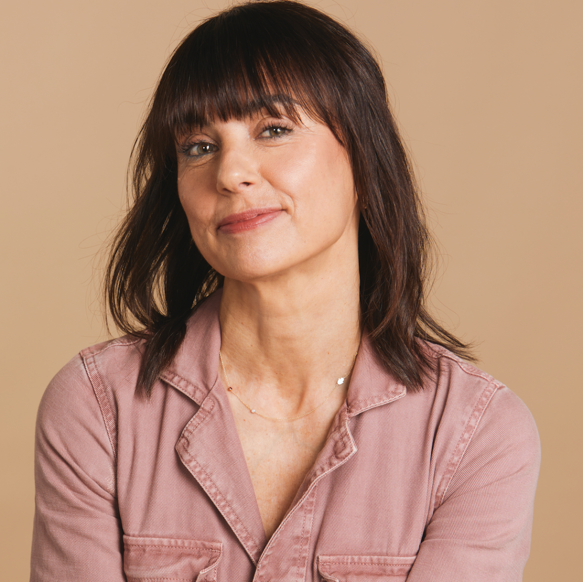 A woman wearing a Custom Mixed Gold Necklace with two birthstones and a hamsa charm, styled in a pink shirt against a beige background. 