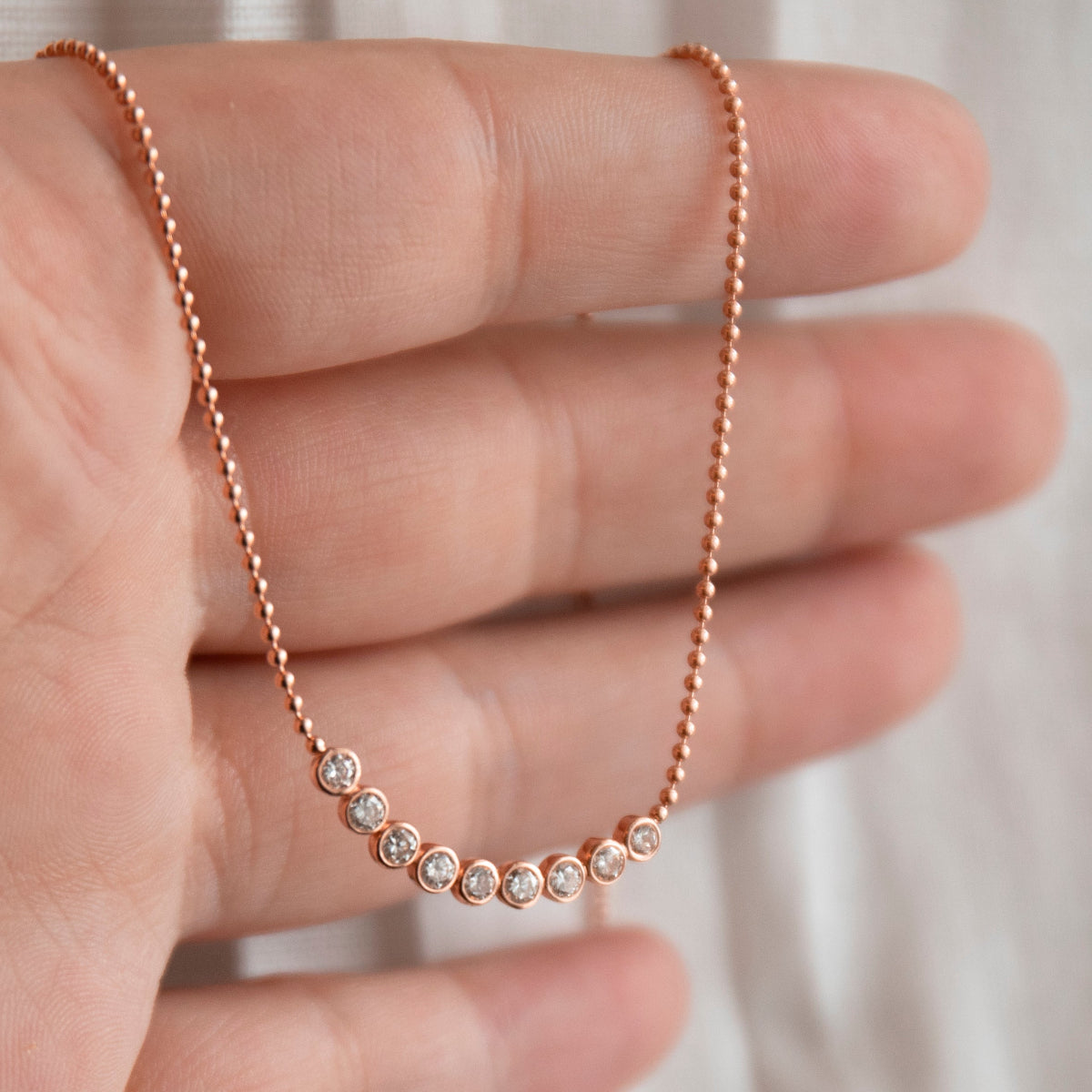 A hand holding a rose gold Diamond Arc Layering Necklace against a off-white background. 