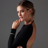A woman wearing a Diamond Cascade Earring, a Bouquet Ring, a Love Set Match Bracelet and a Down the Line Bracelet, styled in a backless black top with blonde hair tied back against a grey background.