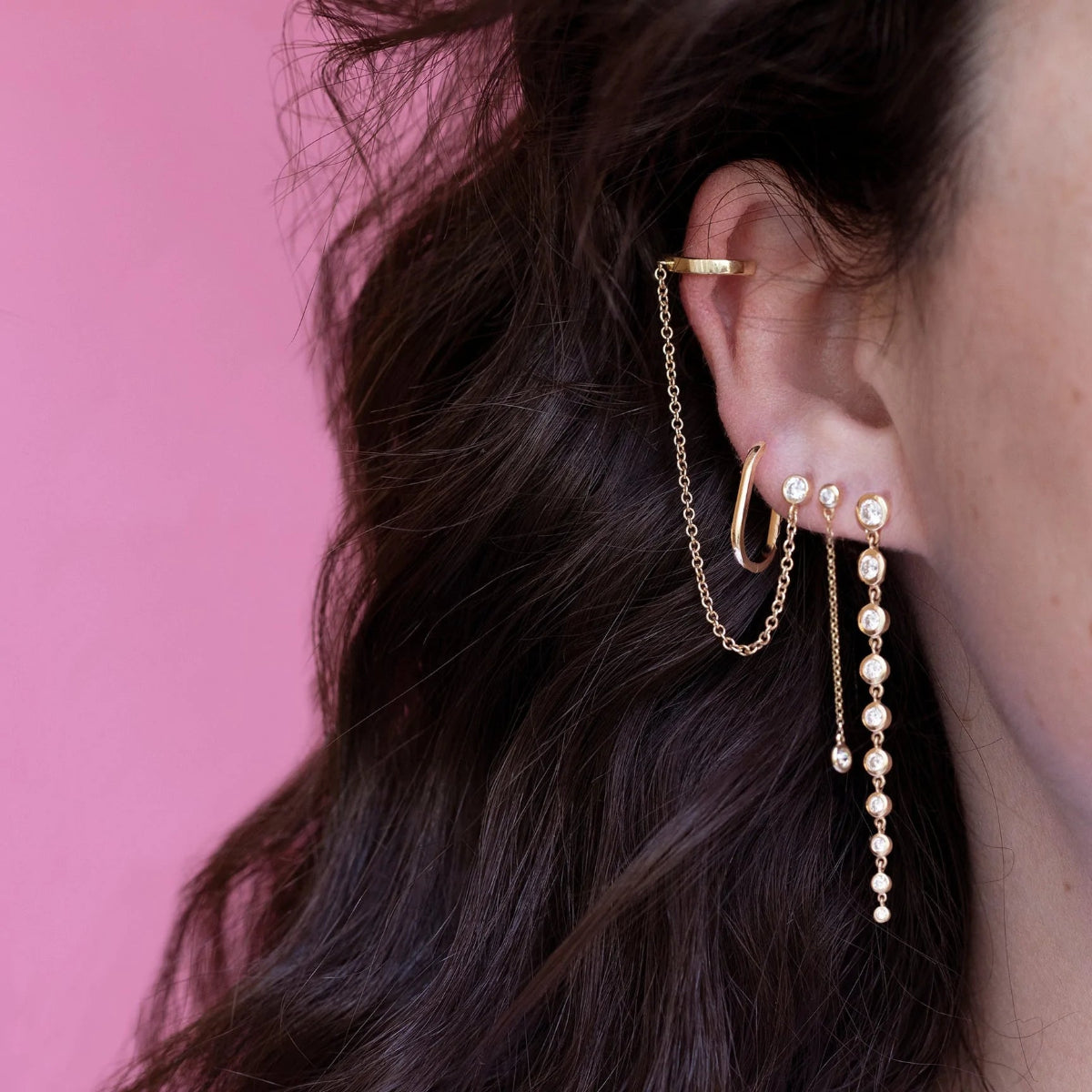 Close-up of a Coupling Ear Cuff + Stud, Oval Hoop, Droplet Diamond Earring and Diamond Cascade Earring in a brunette woman's ear against a pink background. 