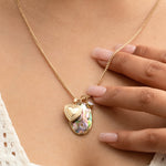 Close-up of a person's décolletage wearing a Round Curb Chain with a Heart Locket Charm, a Large Abalone Cabochon Charm and an Opal Dangle Charm, held by their hand, in a cream top.
