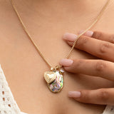 Close-up of a person's décolletage wearing a Round Curb Chain with a Heart Locket Charm, a Large Abalone Cabochon Charm and an Opal Dangle Charm, held by their hand, in a cream top.