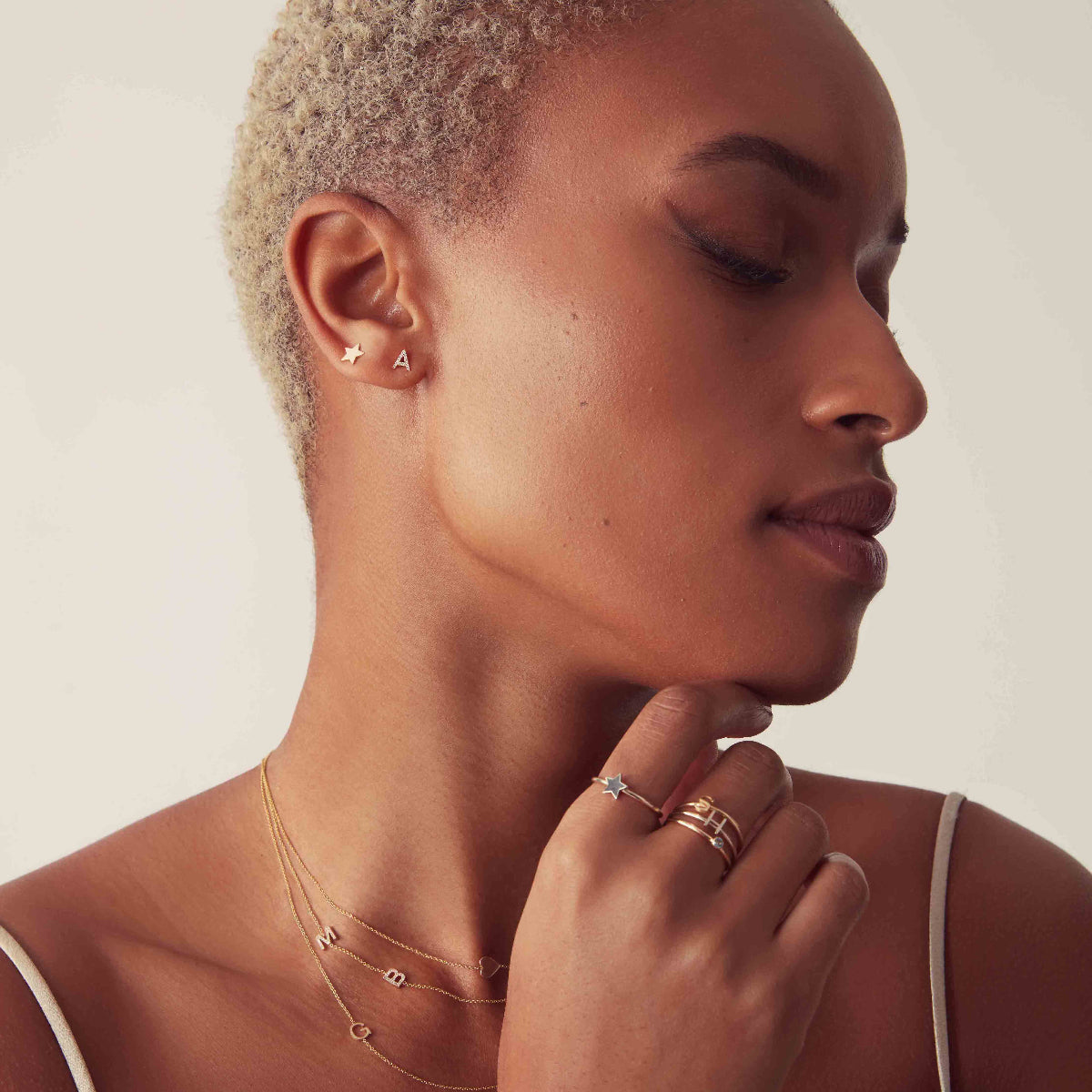 A woman wearing a Heartfelt Stud, a 14K Gold Letter Stud, a 14K Gold Pave Letter Stud, Twinkle Twinkle Ring, a 14K Gold Letter Ring and several letter necklaces,  all in yellow gold, against a white background.   