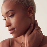A woman with her hand on her neck, wearing three earrings – a pavé letter M stud, a threader earring with a gold heart stud and a gold letter B stud, along with various rings and necklaces.