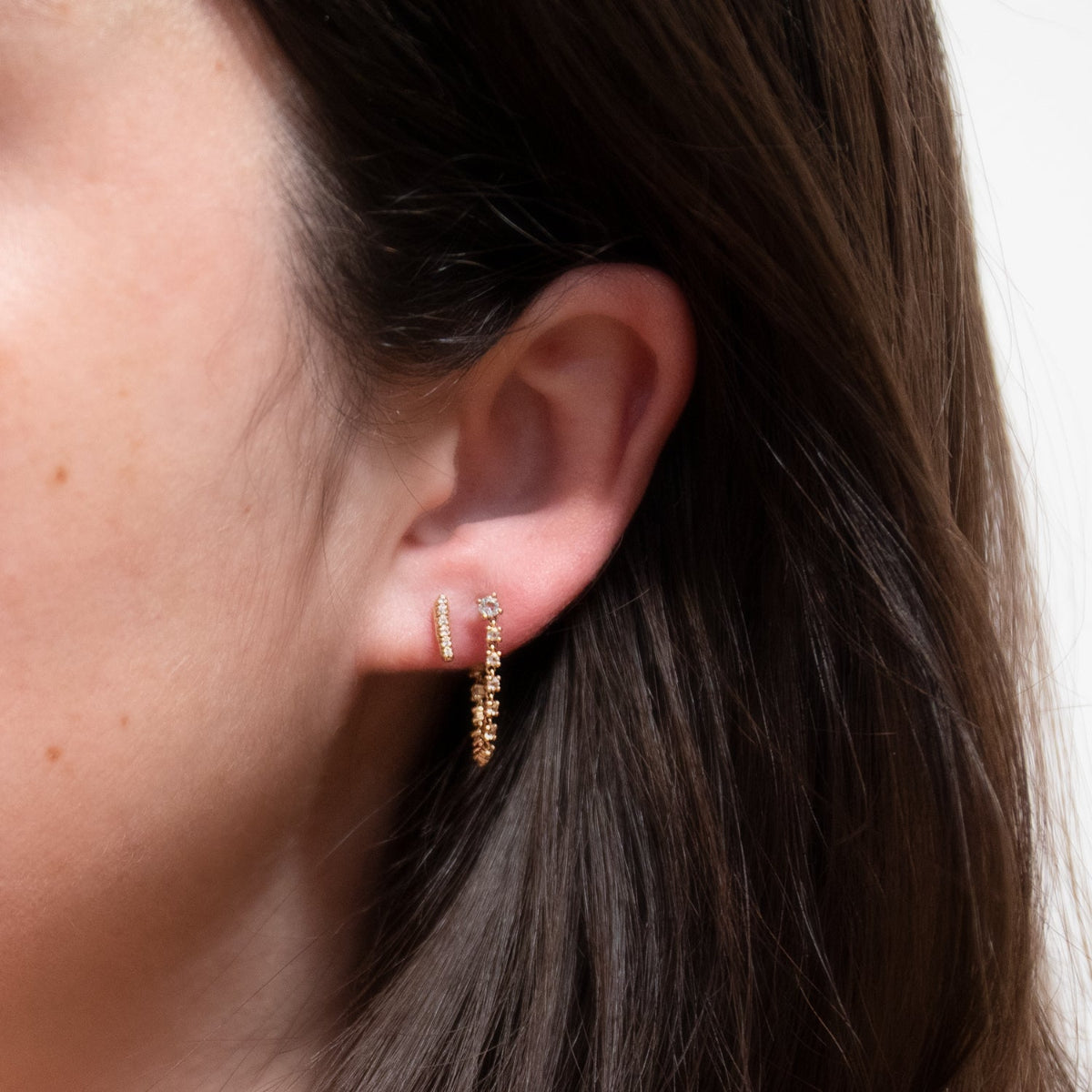 Close-up of woman's ear wearing a White Topaz Chain Earring and a Pave Huggie against a blurred background. 