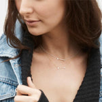 A brunette woman wearing two white gold
Straight as an Arrow Necklaces with a black knit and a light blue denim jacket, against a white background.