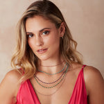 A blonde woman wearing several Bouquet Necklaces (Peony, Bluebell, Thistle, Clover and Sage), yellow gold hoop earrings and a dark pink top against a neutral background. 