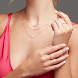 Close-up of a woman’s décolletage wearing the Sweetheart Necklace with pink sapphire, a Quintet Layering Necklace, and a Diamond Arc Layering Necklace. Her hands feature an Open Diamond Ring and pavé diamond rings, styled with a dark pink top against a grey background.