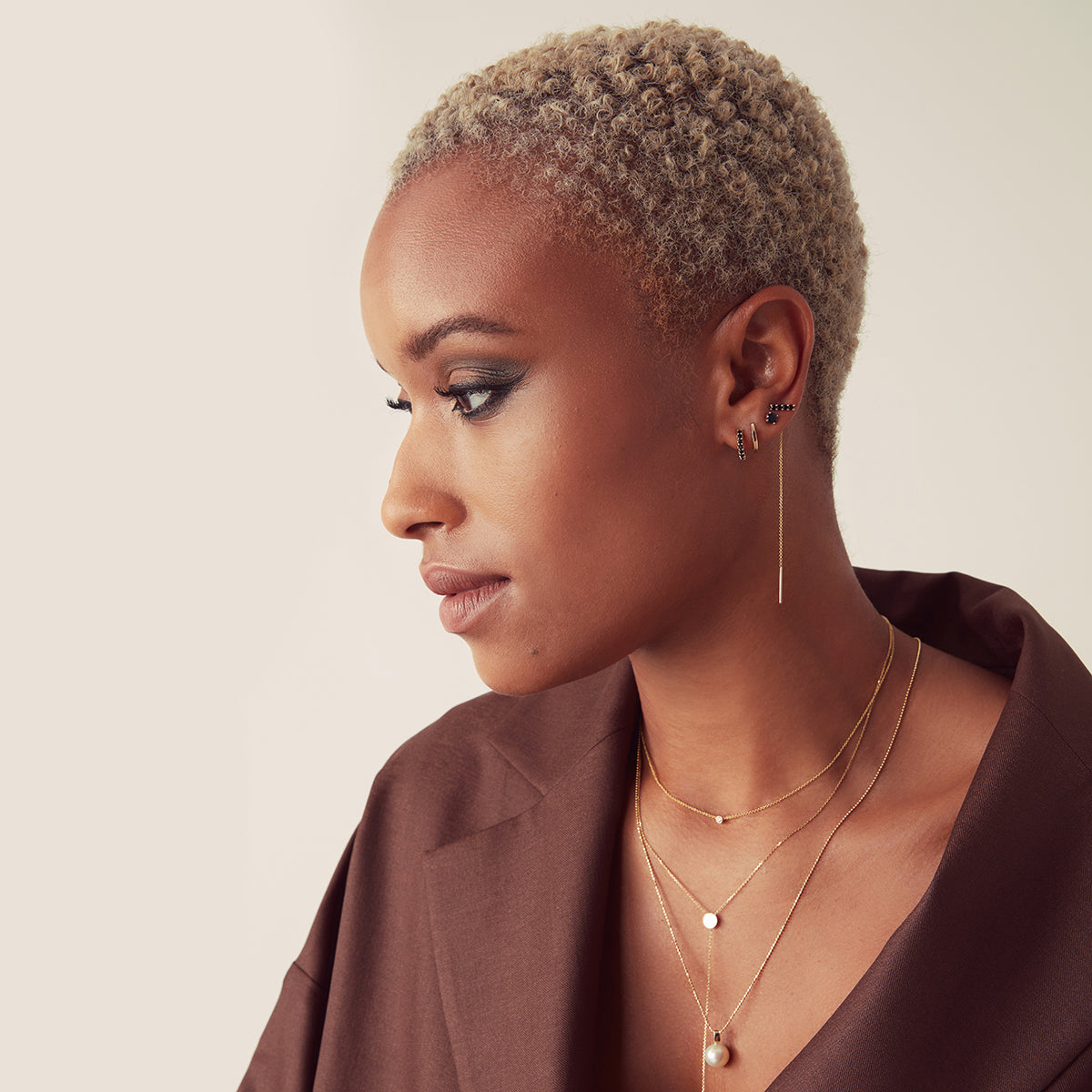 A woman with short blonde hair wearing a Single Diamond Layering Necklace, a Gold Lariat Necklace, a Perla Necklace, an Alli Webb Onyx Comfort Stud, an Onyx Threader, an Onyx Huggie, and a Faceted Huggie, styled in a brown top against a neutral background.