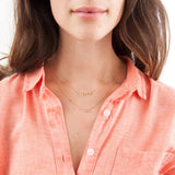A close-up of a brunette woman wearing a FEMME Necklace and an OUI Necklace, with a light orange shirt against a white background.