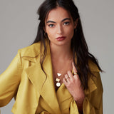 A brunette woman wearing a Geometric Circle Necklace layered with  similar necklaces featuring triangle and rectangle pendants, Sleek & Chic Hoops - Small, a Thick Band and a Tube Ring, all in yellow gold, with a mustard yellow jacket against a light grey background.