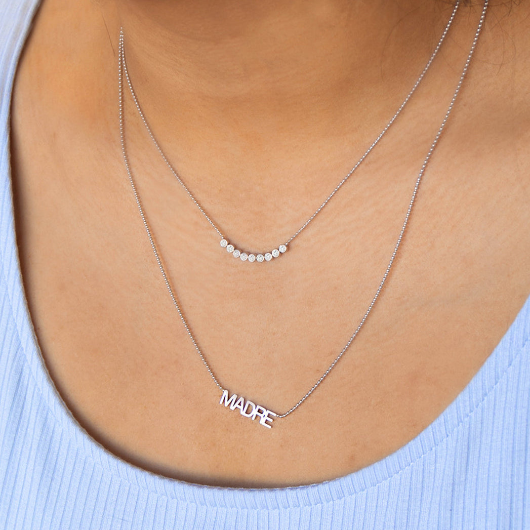 A close-up of a woman's décolletage wearing a Diamond Arc Layering Necklace and a MADRE Necklace, both in white gold, in a light blue top. 
