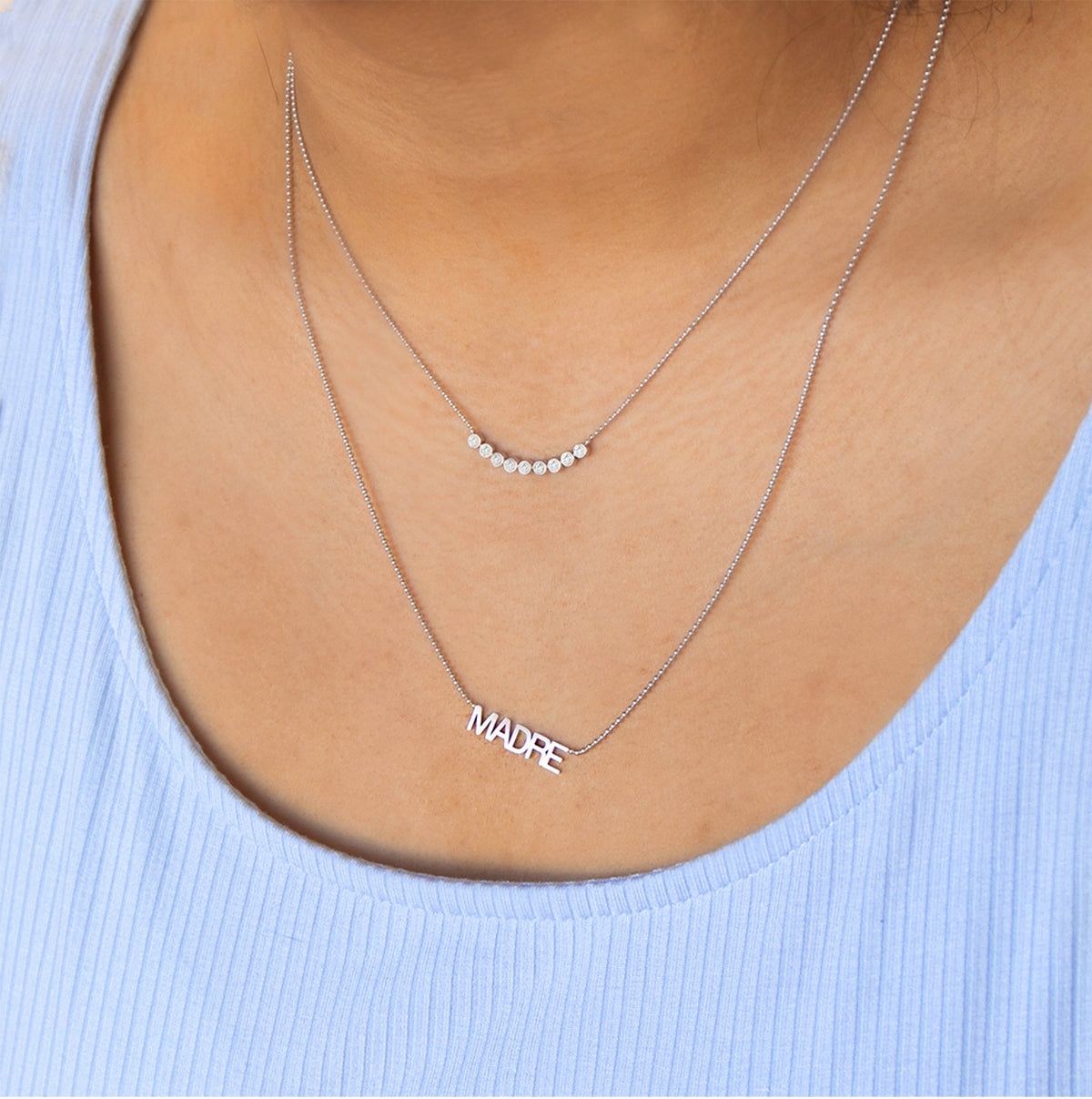 A close-up of a woman's décolletage wearing a Diamond Arc Layering Necklace and a MADRE Necklace, both in white gold, in a light blue top. 