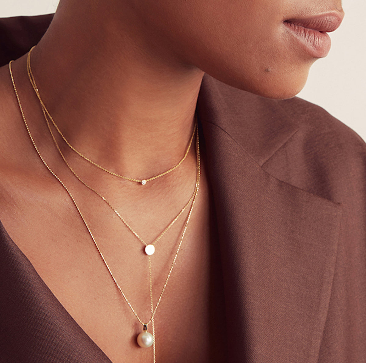 Close-up of a woman's neck wearing a yellow gold Diamond Layering Necklace, a Gold Lariat Necklace and a Perla Necklace, styled in a brown blazer. 