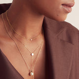 Close-up of a woman's neck wearing a yellow gold Diamond Layering Necklace, a Gold Lariat Necklace and a Perla Necklace, styled in a brown blazer. 