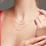 Close-up of a neck wearing The Sweetheart Necklace - Pink Sapphire, Quintet Diamond Layering Necklace and a Diamond Arc Layering Necklace in yellow gold against a grey background. 