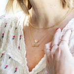 A woman's décolletage wearing an Omega Chain, a Lip Pendant and a BISOUS Necklace, all in yellow gold, with a white top with pink flowers and her short blonde hair visible. 