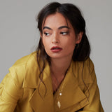 A brunette woman wearing a 20th Anniversary Leaf Necklace, a Perla Necklace and Sleek & Chic Hoops - Small, all in yellow gold, with a yellow mustard jacket against a light grey background. 
