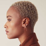 A More Love Stud, Tres Pearl Earring, 14K Gold Charm Threader Earring and Pave Double Ear Cuff worn by a woman with short blonde hair showing her side profile against a light grey background. 