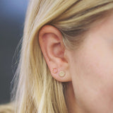 Close-up of an Open Eclipse Earring - Mini and a Solid Eclipse Earring - Mini in yellow gold in a blonde woman's ear against a blue background. 