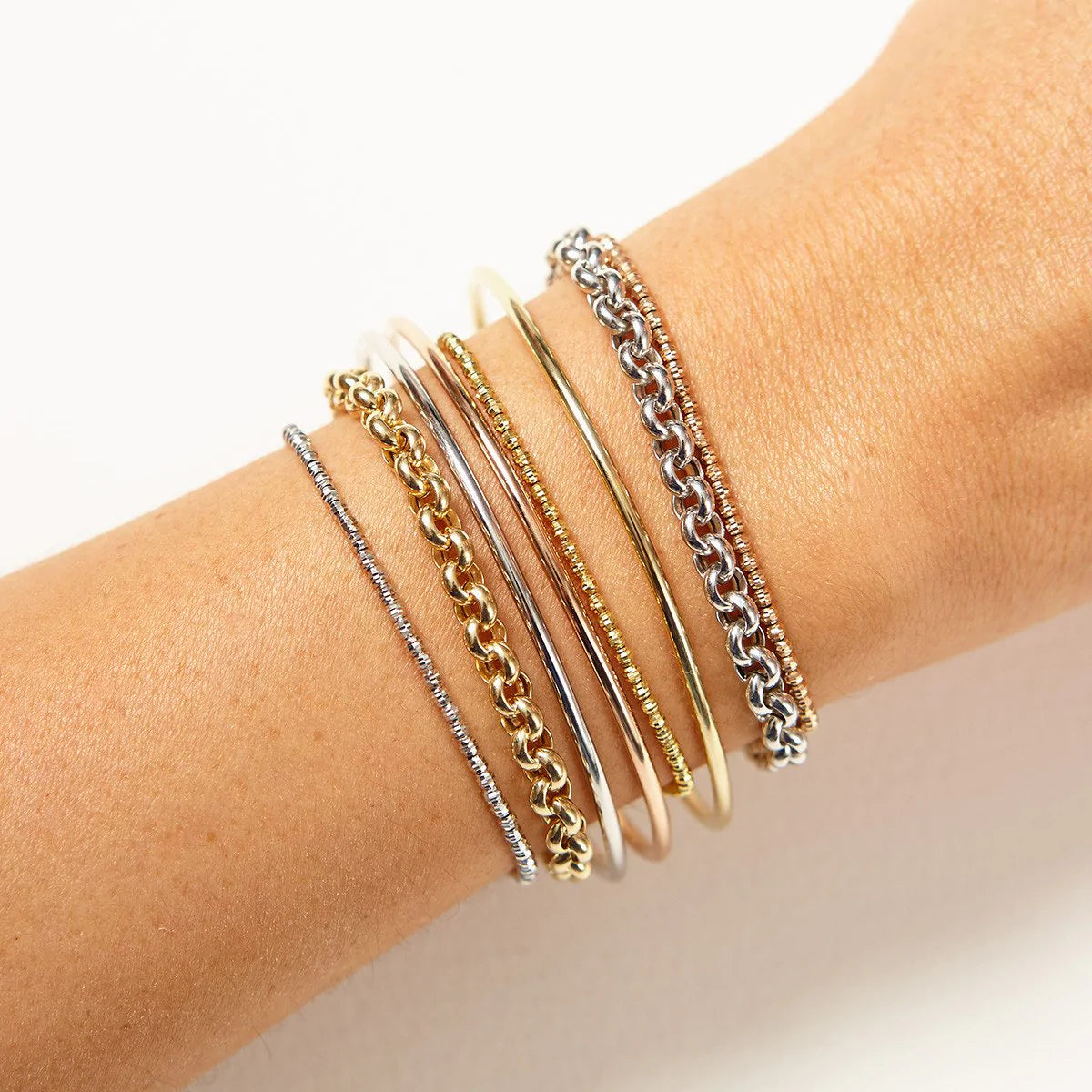 A close-up of an arm wearing mixed metal bracelets: two Rolo Bracelets, three Diamond Cut Bracelets and three Simple Bangles against a white background.