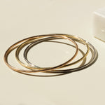 Three Simple Bangles, one in yellow gold, one in rose gold and one in white gold, stacked on a light beige surface. 