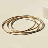 Three Simple Bangles, one in yellow gold, one in rose gold and one in white gold, stacked on a light beige surface. 