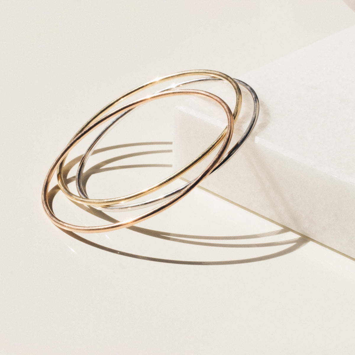 Three Simple Bangles, one in yellow gold, one in rose gold and one in white gold, against an off-white background.