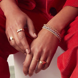 A pair of hands wearing a yellow gold Thick Band, Tres Pearl Ring,  Geo Stacking Ring Square, Delicate Beaded Band, Delicate Twist Band and two Tube Rings, along with mixed metal Rolo Bracelets and Diamond Cut Bracelets, styled in red corduroy clothing. 