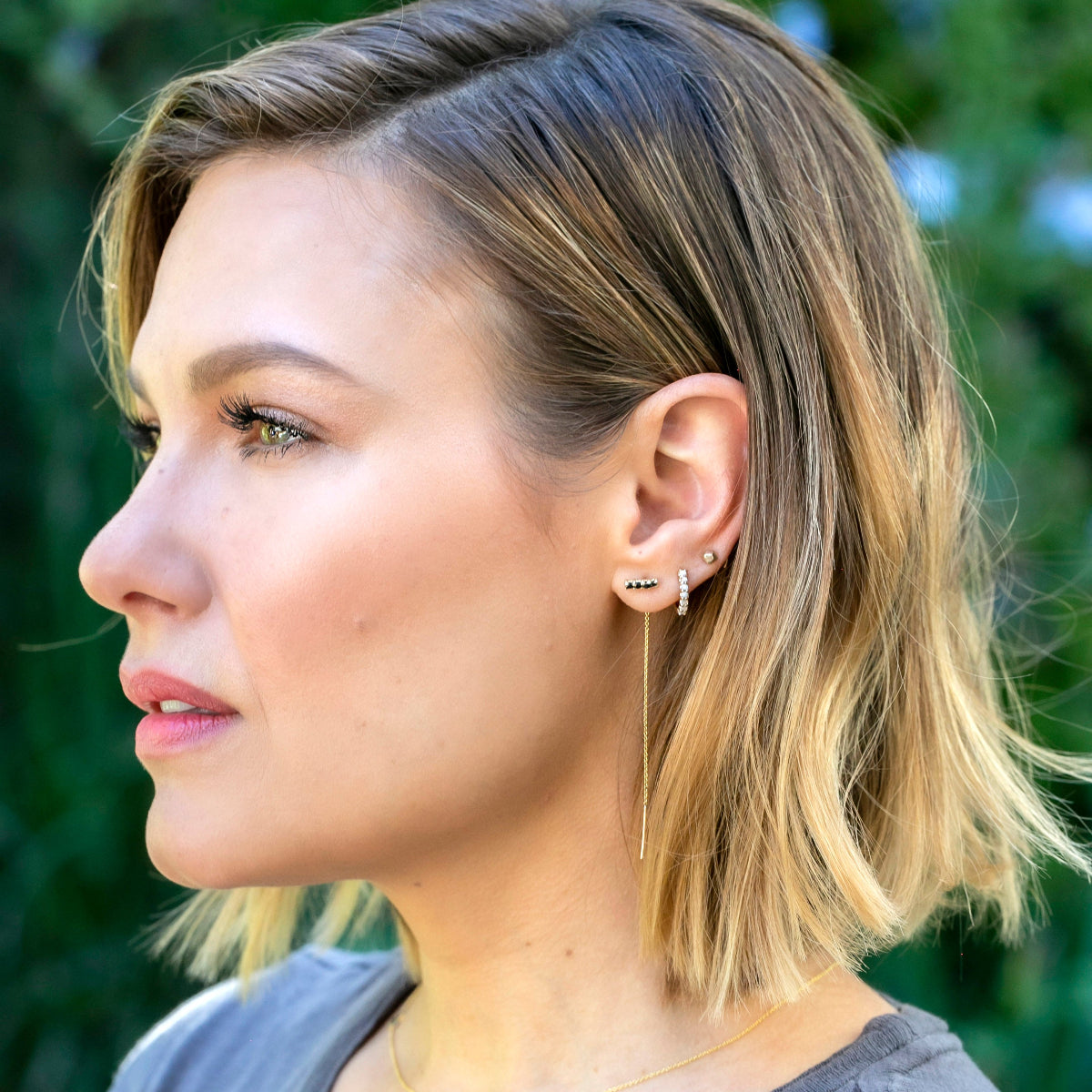 A woman wearing an Alli Webb Onyx Threader, Opal Huggie and a Faceted Comfort Stud, with blonde hair and a grey top, against a green outdoors background. 