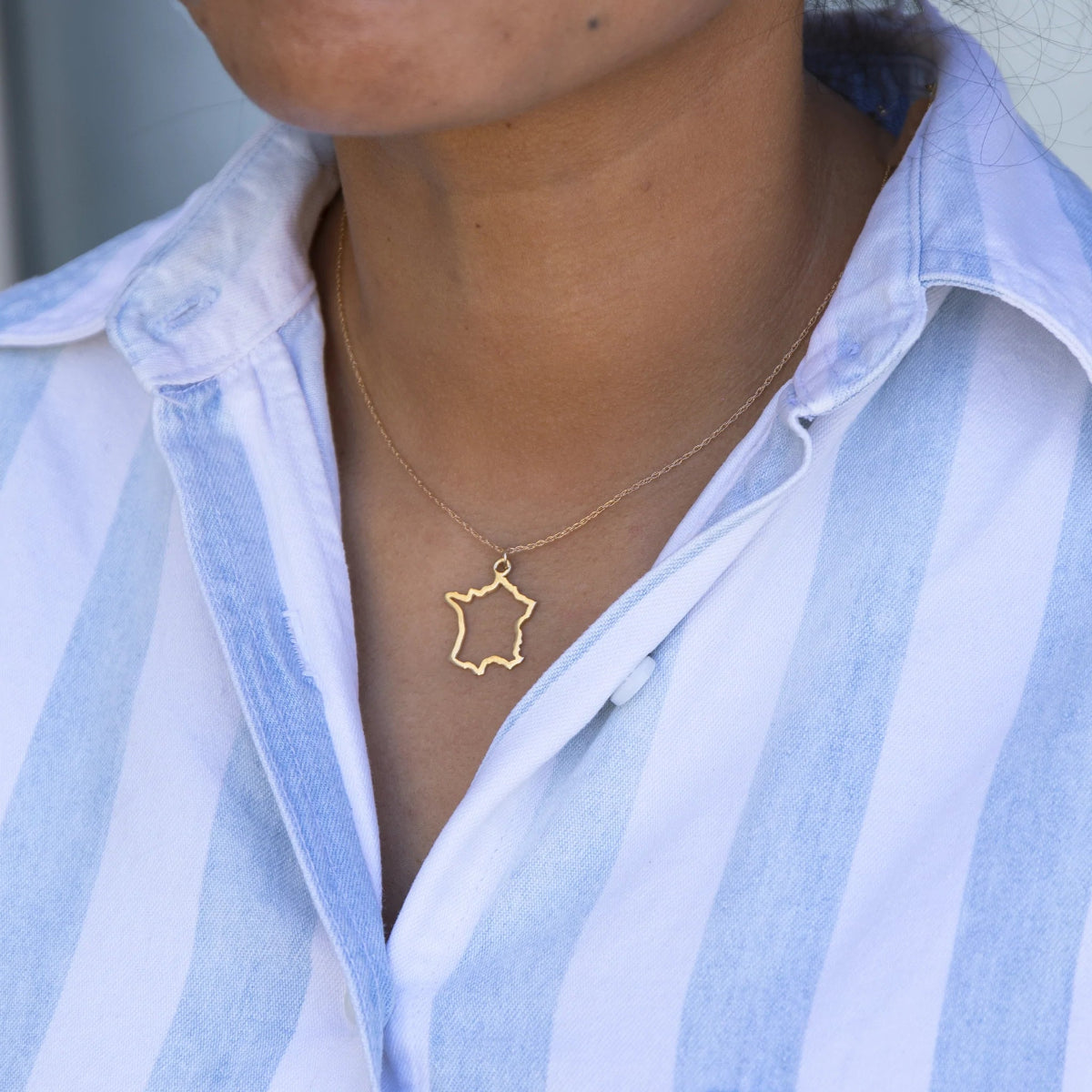 Close-up of a person's neck wearing a France necklace in yellow gold with a blue and white striped shirt against a light grey blurred background. 