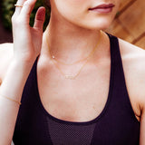 A close-up of a woman's décolletage wearing a RESIST Necklace and a Custom Gold Necklace 1 Letter (X) and a dark purple top. 
