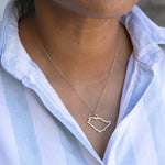 Close-up of a person's neck wearing a Saudi Arabia necklace in white gold with a blue and white striped shirt against a light grey blurred background.