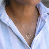 Close-up of a person's neck wearing a Saudi Arabia necklace in white gold with a blue and white striped shirt against a light grey blurred background.