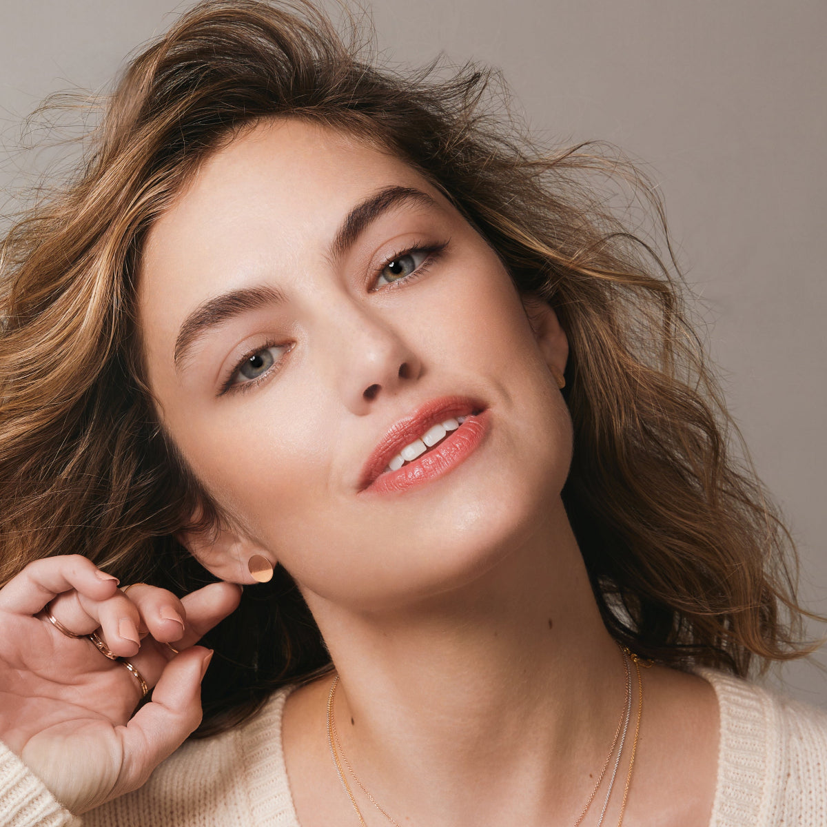 A woman wearing a Solid Eclipse Earring - Small and several gold rings with a cream knit against a light grey background.