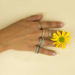A hand wearing several Bouquet Rings in various colours holding a yellow flower against a light yellow background. 
