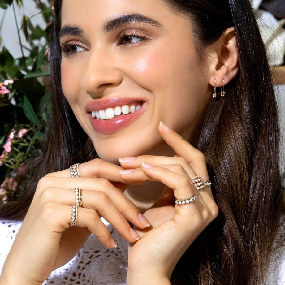 A woman wearing multiple Bouquet Rings, a Gold Safety Pin Earring and Pave Safety Pin Earring, with a white crochet top and brown hair, against a floral background. 