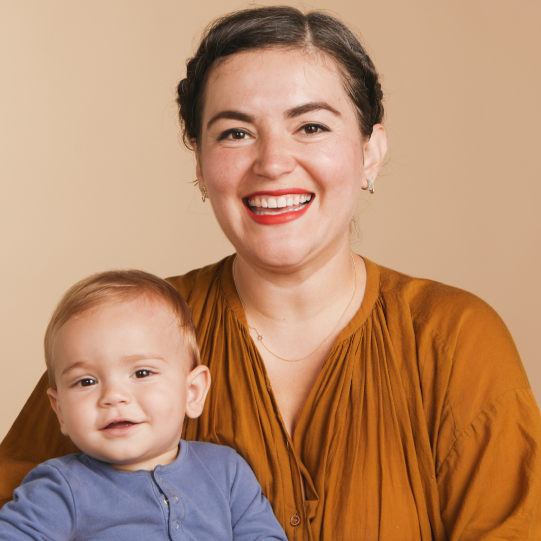 A woman wearing a 14k Gold Asymmetrical Charm Necklace - Star of David and various earrings, styled in a burnt orange top, holding a baby wearing a blue top, against a neutral background.