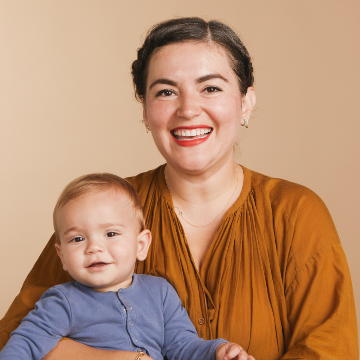 A woman wearing a 14k Gold Asymmetrical Charm Necklace - Star of David and various earrings, styled in a burnt orange top, holding a baby wearing a blue top, against a neutral background. 
