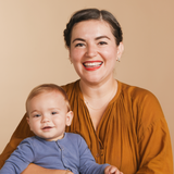 A woman wearing a 14k Gold Asymmetrical Charm Necklace - Star of David and various earrings, styled in a burnt orange top, holding a baby wearing a blue top, against a neutral background. 
